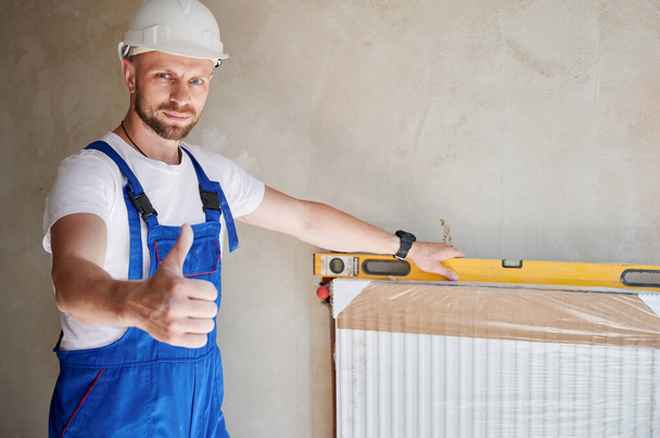 Male worker in construction helmet looking at camera and showing approval gesture while installing heating radiator in apartment under renovation. Man in work overalls using spirit level ruler tool. - Photo, Image