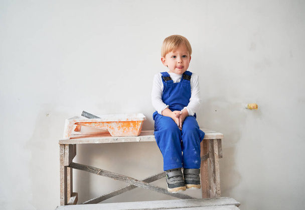 Cute kid construction worker sitting on wooden table against white wall in apartment under renovation. Cheerful little boy wearing safety helmet and work overalls while playing at home. - Φωτογραφία, εικόνα