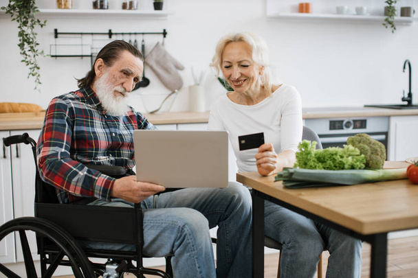 Old Caucasian couple charming wife and bearded man in wheelchair paying for food with credit card using laptop against background of spacious kitchen. - Foto, afbeelding