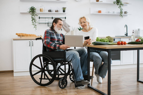 Old Caucasian couple charming wife and bearded man in wheelchair paying for food with credit card using laptop against background of spacious kitchen. - Fotó, kép