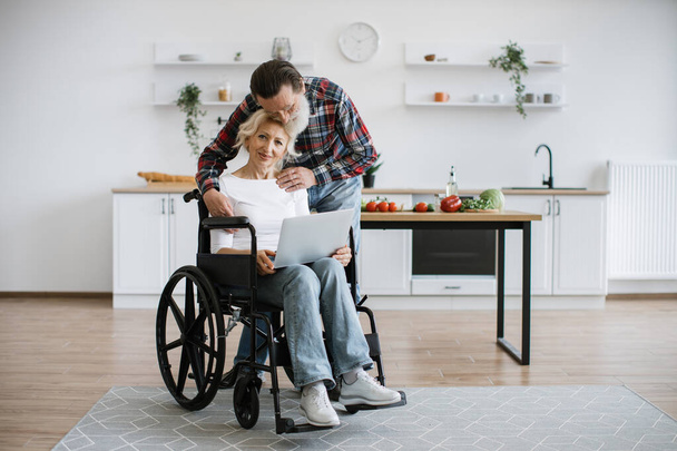 Charming old couple of woman in wheelchair with laptop and husband hugging her on background of bright modern kitchen, looking at camera. - Zdjęcie, obraz