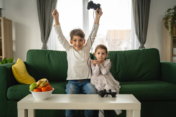 Két gyermek kis kaukázusi testvér és testvér boldog gyerekek testvérek fiú és lány játszik videojáték konzol segítségével joystick vagy vezérlő, miközben otthon ül valós emberek családi szabadidős koncepció - Fotó, kép