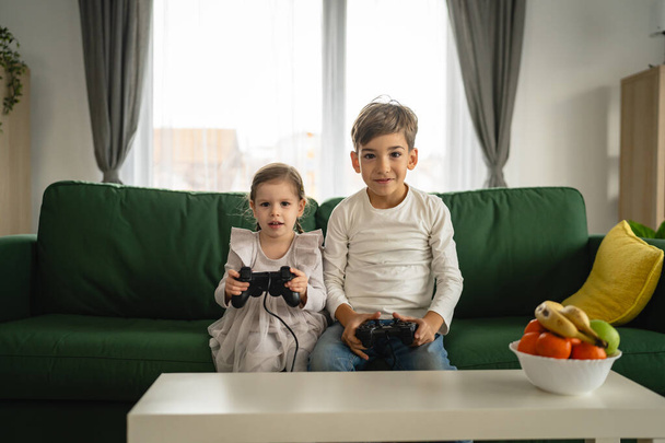 Duas crianças pequeno caucasiano irmão e irmã feliz crianças irmãos menino e menina jogando console de vídeo game usando joystick ou controlador enquanto sentado em casa pessoas reais conceito de lazer familiar - Foto, Imagem