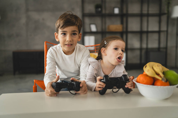 Δύο παιδιά μικρός καυκάσιος αδελφός και αδελφή ευτυχισμένα παιδιά αδέλφια αγόρι και κορίτσι που παίζουν κονσόλα βιντεοπαιχνιδιών χρησιμοποιώντας χειριστήριο ή χειριστήριο ενώ κάθονται στο σπίτι πραγματικοί άνθρωποι έννοια οικογενειακή αναψυχή - Φωτογραφία, εικόνα