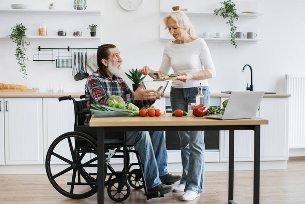 Senior couple, man in wheelchair and elderly woman, making healthy salad. Male with gray beard holding bowl while his partner pours chopped vegetables. - Fotó, kép