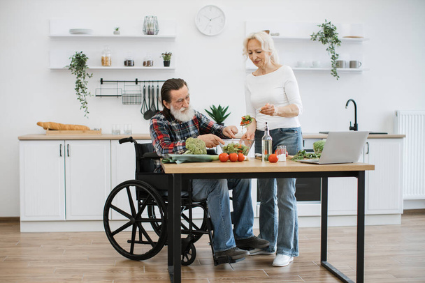 Old couple with disability spends free time cooking breakfast in modern light kitchen. Seniors husband in wheelchair and wife mixing chopped vegetables in bowl while prepare delicious healthy salad. - Foto, Bild