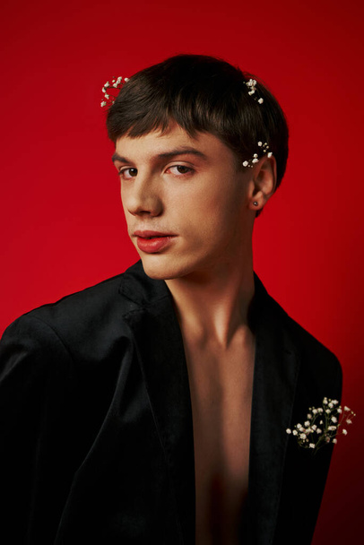 красивый молодой человек в стильном наряде с цветами в волосах, смотрящий в камеру на красном фоне - Фото, изображение