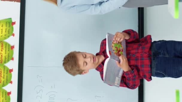 Vertikale Ansicht eines kaukasischen Schülers, der ein Buch in der Hand hält, während er in der Mitte im Klassenzimmer steht. Intelligenter Junge, der seinen Klassenkameraden Texte oder Hausaufgaben vorliest. Bildung im Vorschul- oder Kindergartenalter. - Filmmaterial, Video