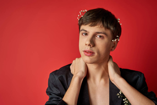 young man in velvet blazer with flowers in hair looking at camera and touching neck on red backdrop - Photo, Image