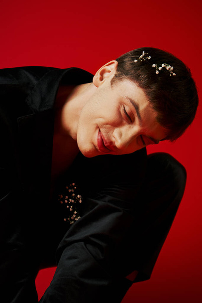cheerful young man in black velvet jacket with flowers in hair smiling on red background, glamour - Photo, Image