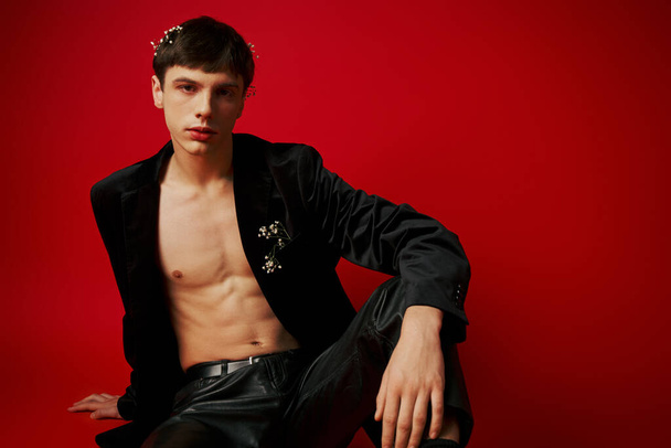 young male model in black attire with flowers in hair and leather pants sitting on red background - Photo, Image