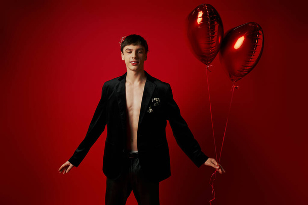 joven feliz y guapo en traje negro sosteniendo globos en forma de corazón sobre fondo rojo - Foto, Imagen