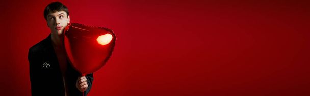 jeune homme confus en veste de velours tenant ballon en forme de coeur et regardant loin sur rouge, bannière - Photo, image