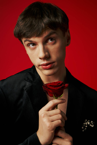 ρομαντικός και ονειροπόλος νεαρός άνδρας με βελούδινο σακάκι που κρατά τριαντάφυλλο στα χέρια του σε κόκκινο φόντο, φλερτάρει - Φωτογραφία, εικόνα