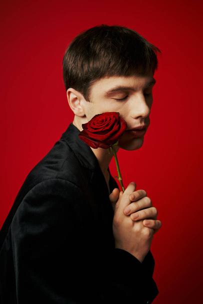 retrato de tipo romántico con los ojos cerrados sosteniendo rosa cerca de la mejilla sobre fondo rojo, flor - Foto, imagen