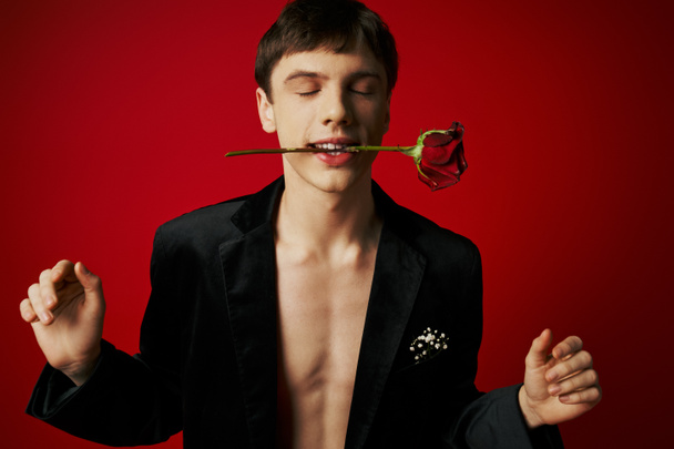 portrait of young man with closed eyes holding rose in teeth and smiling on red background, romance - Photo, Image