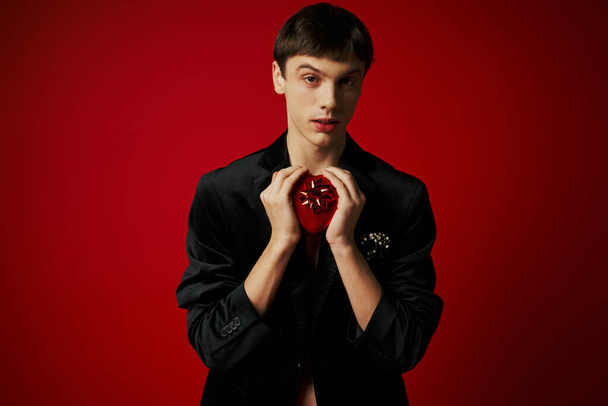 Elinde kalp şeklinde bir hediye olan romantik adam kırmızı arka plandaki kameraya bakıyor. Sevgililer Günü 'nde. - Fotoğraf, Görsel
