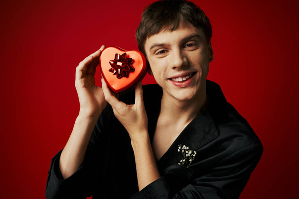 vrolijke jongeman in fluwelen blazer met hartvormig cadeau op rode achtergrond, Valentijnsdag - Foto, afbeelding