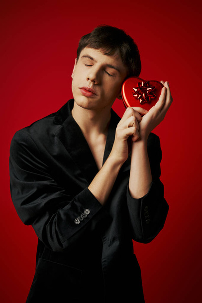 Kırmızı arka planda kadife ceketli, kalp şeklinde hediyeler taşıyan duygusal bir adam, Sevgililer Günü. - Fotoğraf, Görsel