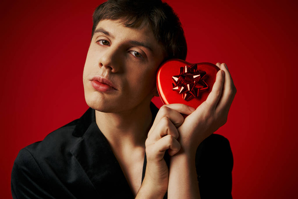 hombre sentimental en chaqueta de terciopelo con caja de regalo en forma de corazón sobre fondo rojo, día de San Valentín - Foto, imagen