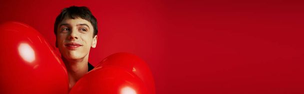 feliz joven sonriendo cerca de globos en forma de corazón sobre fondo rojo, bandera del día de San Valentín - Foto, Imagen