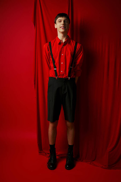 felice giovane uomo in pantaloncini con bretelle e camicia in piedi vicino alla tenda rossa, look alla moda - Foto, immagini