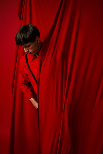 pensive jongeman in levendige shirt met bretels verstopt achter rood gordijn, modieuze look - Foto, afbeelding