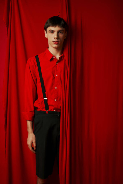 νεαρός άνδρας με ζωντανό πουκάμισο και σορτς με τιράντες που κρύβονται πίσω από την κόκκινη κουρτίνα, μοντέρνα εμφάνιση - Φωτογραφία, εικόνα