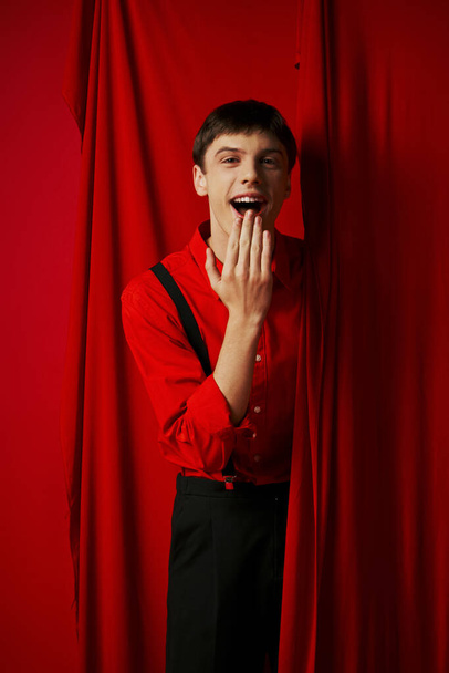asombrado joven en tirantes sonriendo y cubriendo la boca cerca de rojo vibrante cortina, alegría - Foto, Imagen