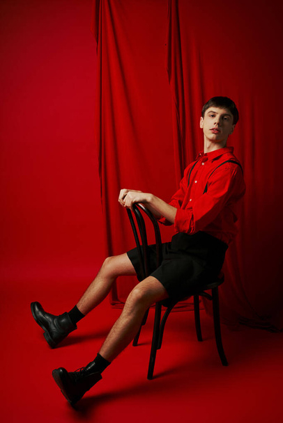 jonge man in shirt en zwarte shorts zitten op stoel met zelfverzekerde pose naast rood gordijn - Foto, afbeelding