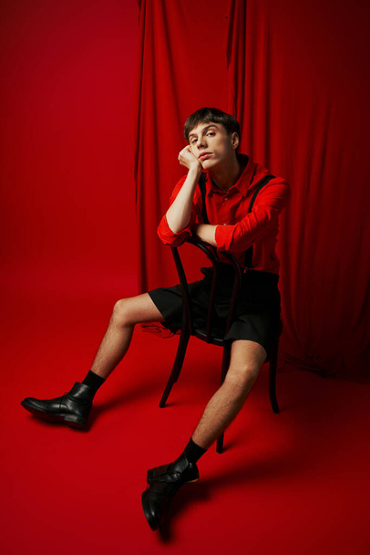 bored young man in shirt and black shorts sitting on chair with relaxed pose next to red curtain - Photo, Image