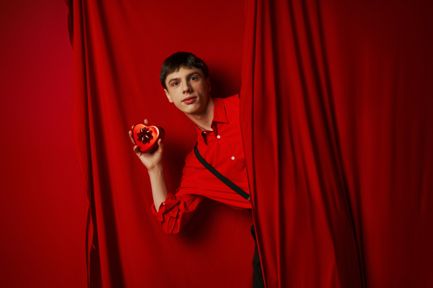 giovane uomo in camicia rossa con bretelle in possesso di scatola regalo a forma di cuore vicino alla tenda, 14 febbraio - Foto, immagini