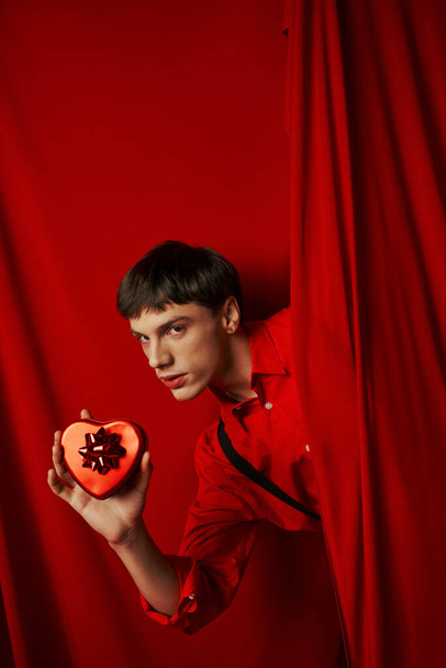 giovane uomo in camicia rossa con bretelle in possesso di cuore a forma di regalo vicino alla tenda, San Valentino - Foto, immagini