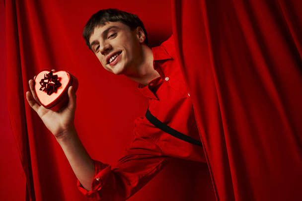 hombre feliz en camisa roja con tirantes con corazón en forma de regalo cerca de la cortina, día de San Valentín - Foto, imagen