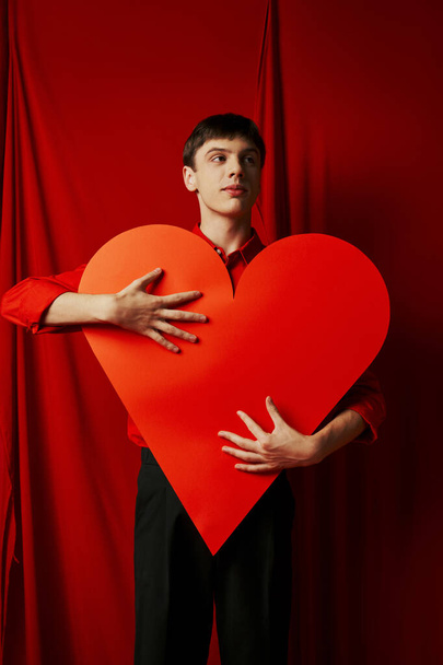 ψηλός νεαρός άνδρας με μαύρο σορτς αγκαλιάζει μεγάλη καρδιά κοπεί σε κόκκινο φόντο, Ημέρα του Αγίου Βαλεντίνου - Φωτογραφία, εικόνα