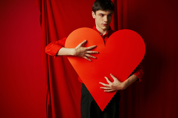 junger Mann in schwarzen Shorts umarmt großen herzförmigen Karton auf rotem Hintergrund, Valentinstag - Foto, Bild