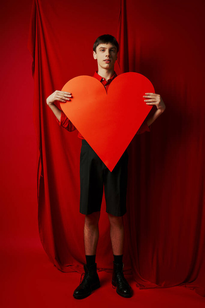 full length of tall man embracing large heart shaped carton on red background, Valentines day - Photo, Image