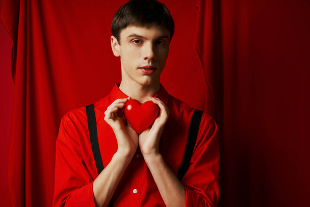 Молодой человек держит маленькое сердце в руках и смотрит на камеру на красном фоне, 14 февраля - Фото, изображение