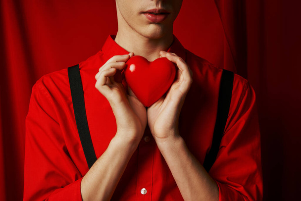 обрезанный вид молодого человека с маленьким сердцем в руках на красном фоне, День святого Валентина концепции - Фото, изображение
