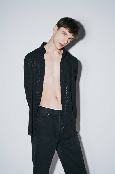 joven guapo en camisa abierta negro brillante posando con las manos detrás de la espalda sobre fondo gris - Foto, Imagen