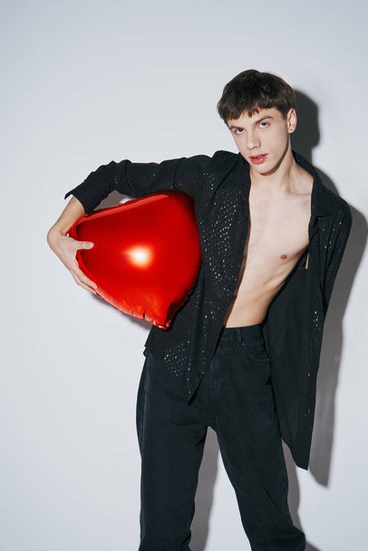 handsome young man in black shiny open shirt holding red balloon on grey background, 14 February - Photo, Image