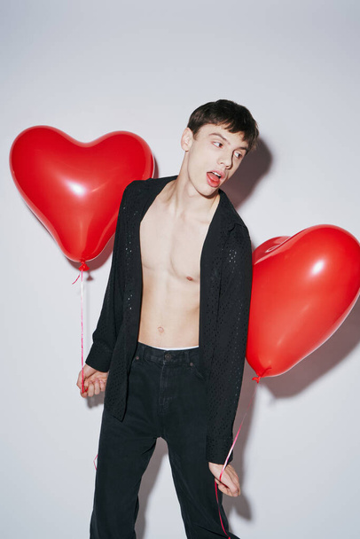 young man in black shirt posing with open mouth and holding red balloons on grey background - Photo, Image