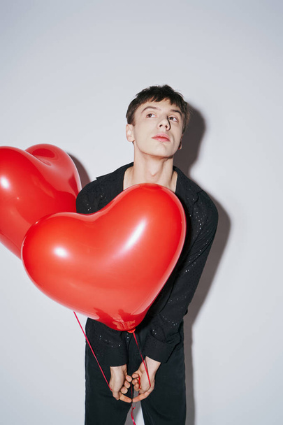młody mężczyzna w czarnej koszuli trzymając czerwone serce w kształcie balonów na szarym tle, Walentynki - Zdjęcie, obraz
