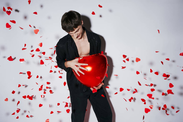 joven en jeans y camisa sosteniendo globo rojo cerca de confeti en forma de corazón sobre fondo gris - Foto, Imagen