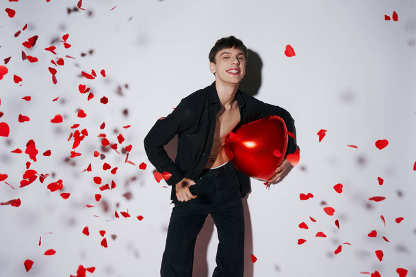 hombre sonriente en jeans y camisa sosteniendo globo rojo cerca de confeti en forma de corazón sobre fondo gris - Foto, imagen