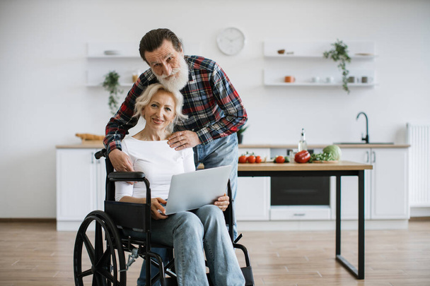 Close up of carming old couple of woman in wheelchair with laptop and husband hugging her on background of bright modern kitchen, looking at camera. - 写真・画像