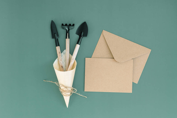 Three garden tools of two spatulas, one rake in a wooden cone and a craft envelope with a leaf lying in the center on a green background with copy space flat lay close-up.Gardening concept,gifts. - Photo, Image