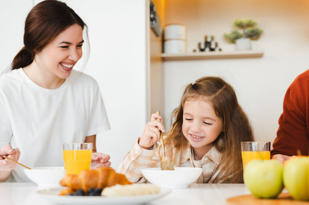 Portrait der schönen jungen Mutter und ihrer kleinen Tochter beim gemeinsamen Frühstück, am Tisch in der Küche sitzend und leckere Gerichte zu sich nehmend. Ernährungskonzept - Foto, Bild