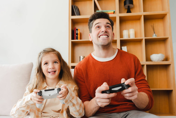 Retrato de homem sorridente, bonito e sua filhinha jogando videogames, segurando joystick, sentado em casa em quarto acolhedor. Conceito de tecnologia online - Foto, Imagem