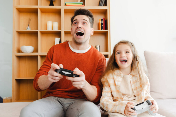 Porträt eines aufgeregten jungen Vaters und seiner kleinen Tochter, die eine Spielkonsole in der Hand halten, Videospiele spielen, zu Hause in einem gemütlichen Zimmer sitzen und Spaß haben. Online-Technologiekonzept - Foto, Bild